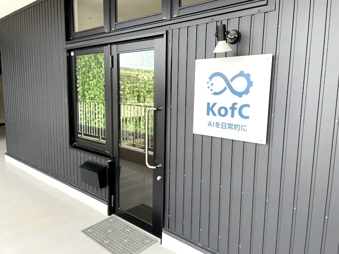 KofC事務所の入口。黒の縦張り外壁とガラス扉、看板に「AIを日常的に」